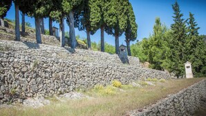 Eine steinerne Stützmauer mit einem kleinen Gebäude darauf, umgeben von Bäumen und Gras.