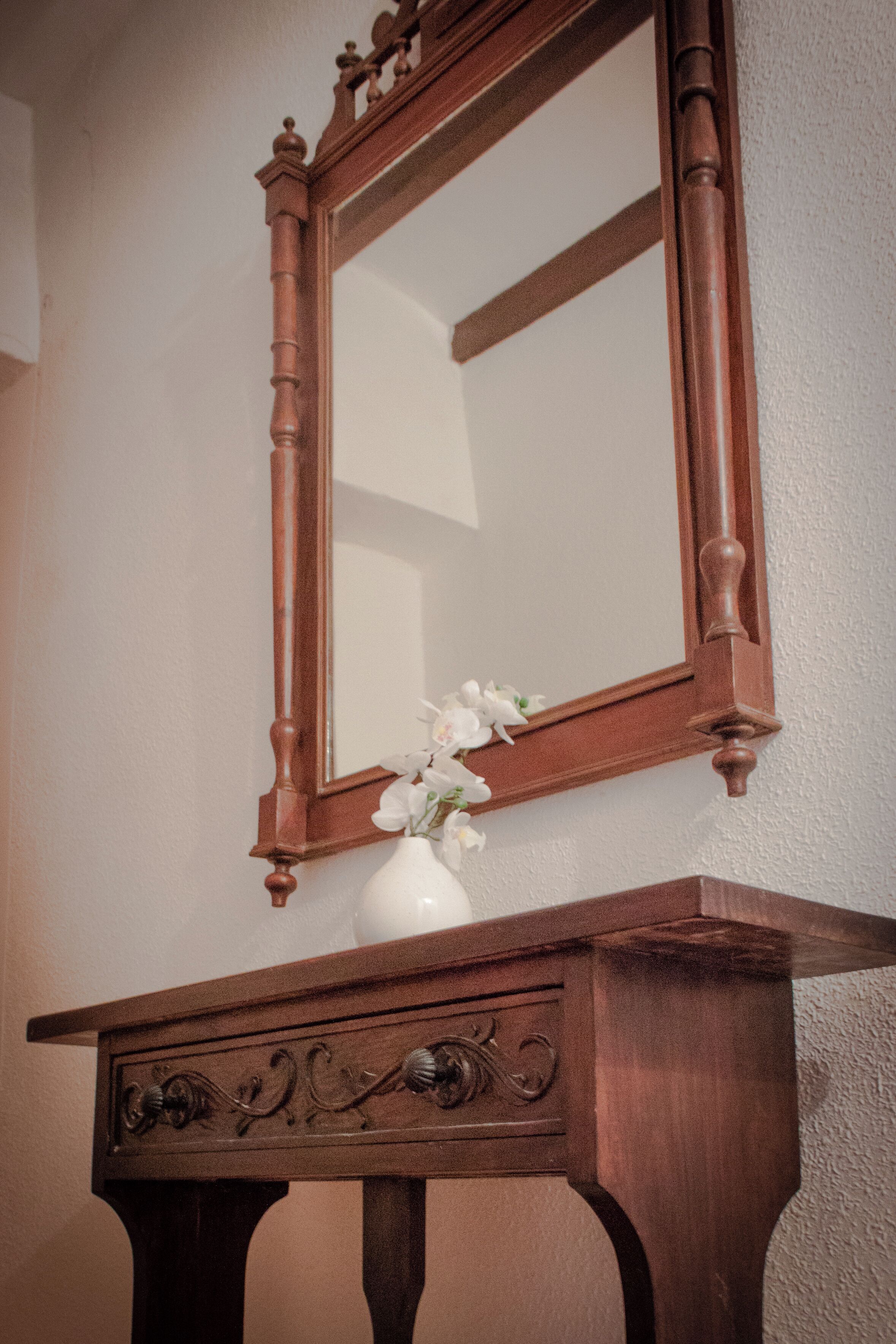 Een houten spiegel met een vaas witte bloemen op een donker houten tafeltje tegen een effen muur.