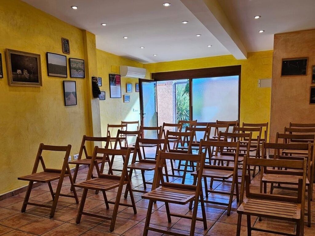 Una habitación con varias sillas de madera dispuestas para sentarse, piso de baldosas y un ventanal que proporciona luz natural.
