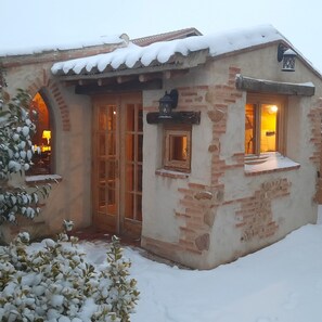 A rustic building with a tiled roof, a wooden door, and a window with a lantern. Snow covers the ground and plants.