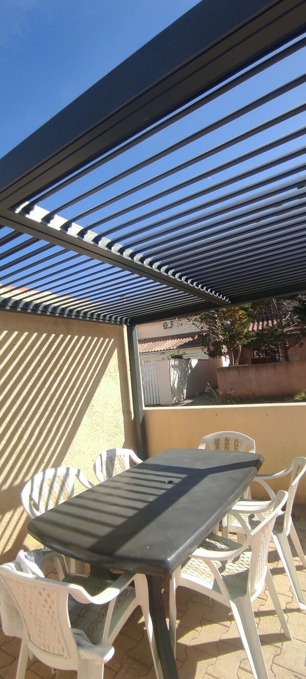 An outdoor patio with a table and chairs, a pergola overhead, and a view of a building in the background.