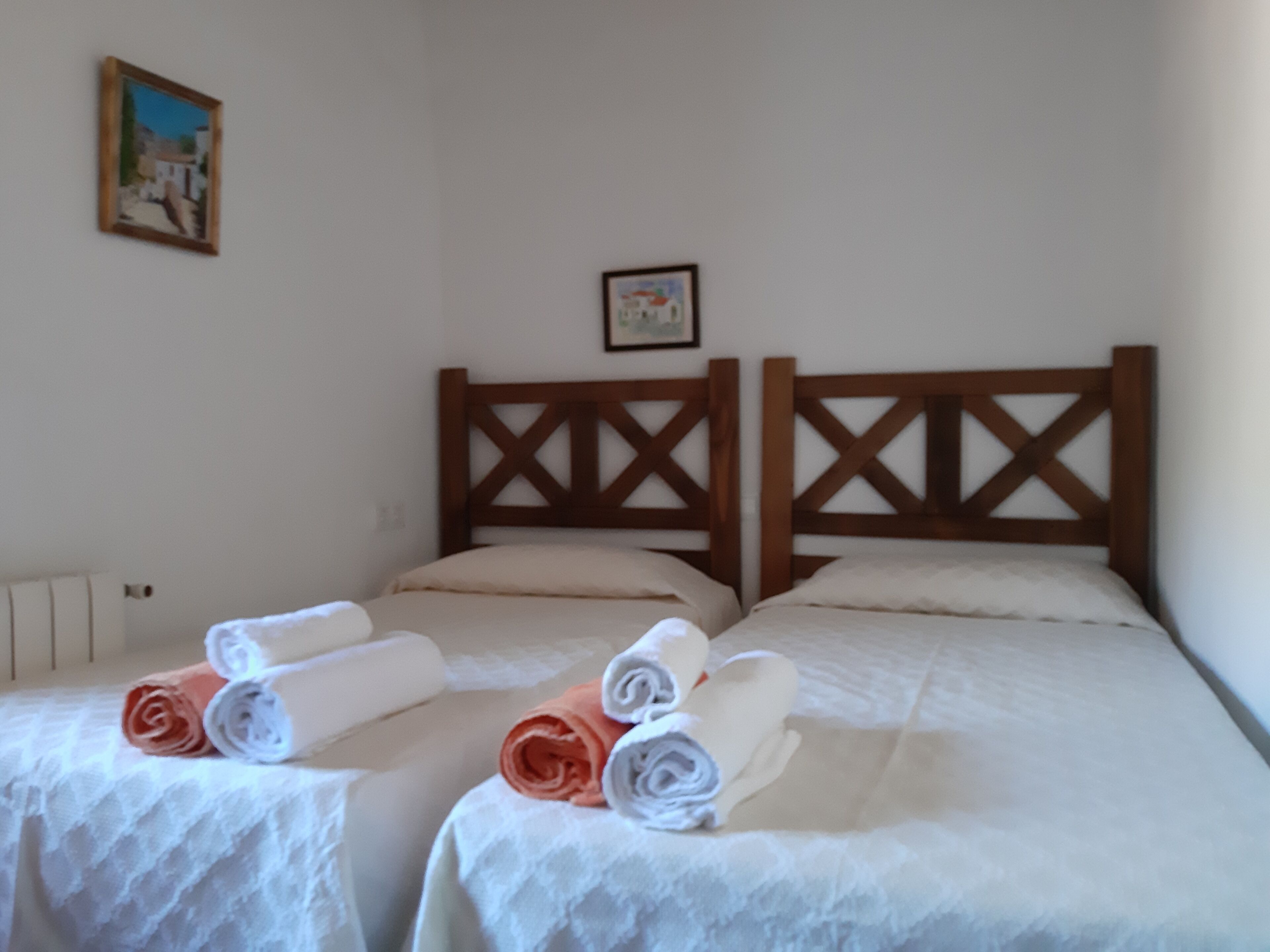 A hotel room with two beds, each with a wooden headboard and white linens. There are rolled towels on the beds.