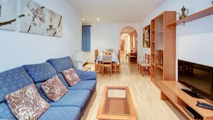 A living room with a blue sofa, a coffee table, a TV, and a dining area with a table and chairs. - P.C. LLEVANT 3 1 Roses (Rosas / Roses)