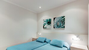A double bed with blue bedding, a bedside table with a lamp, and two wall-mounted artworks. - P.C. LLEVANT 3 1 Roses (Rosas / Roses)