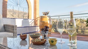 A glass table with a bottle of white wine, two wine glasses, a bowl of olives, a bowl of nuts, and a decorative pot on a balcony with a view of a brick wall and a large window. - Alojamiento Rural El Valle for 6 people (Dúrcal)