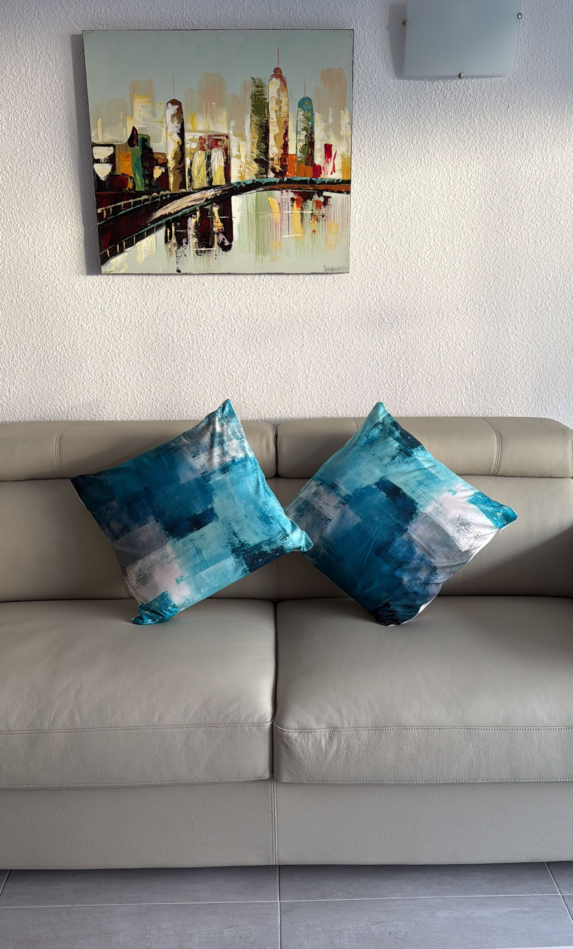 Un divano in pelle beige con due cuscini a motivi blu e un quadro raffigurante un panorama urbano.
