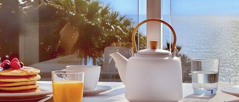 A breakfast setup with pancakes, orange juice, a teapot, and a glass of water on a table with a view of the ocean.
