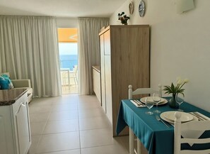 Un comedor con una mesa preparada para dos, vista al océano a través de una ventana y una zona de cocina con un armario blanco.