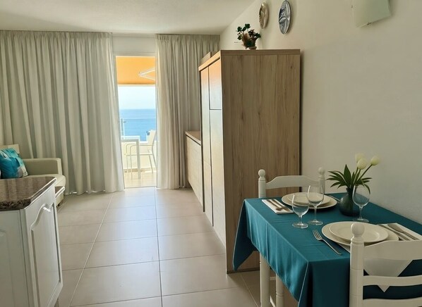 A dining area with a table set for two, a view of the ocean through a window, and a kitchen area with a white cabinet.