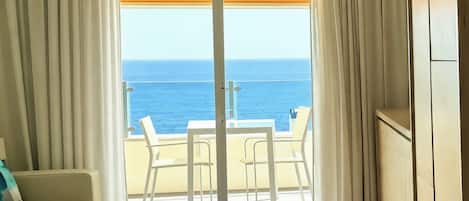 A room with a view of the ocean, a sliding glass door, a table with chairs, and a wooden cabinet.