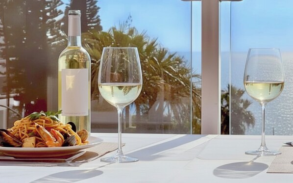 Una mesa con un plato de pasta de mariscos, dos copas de vino y una botella de vino blanco, frente a un paisaje pintoresco.