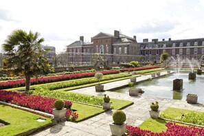 A formal garden with a fountain, neatly trimmed hedges, and a large building in the background.