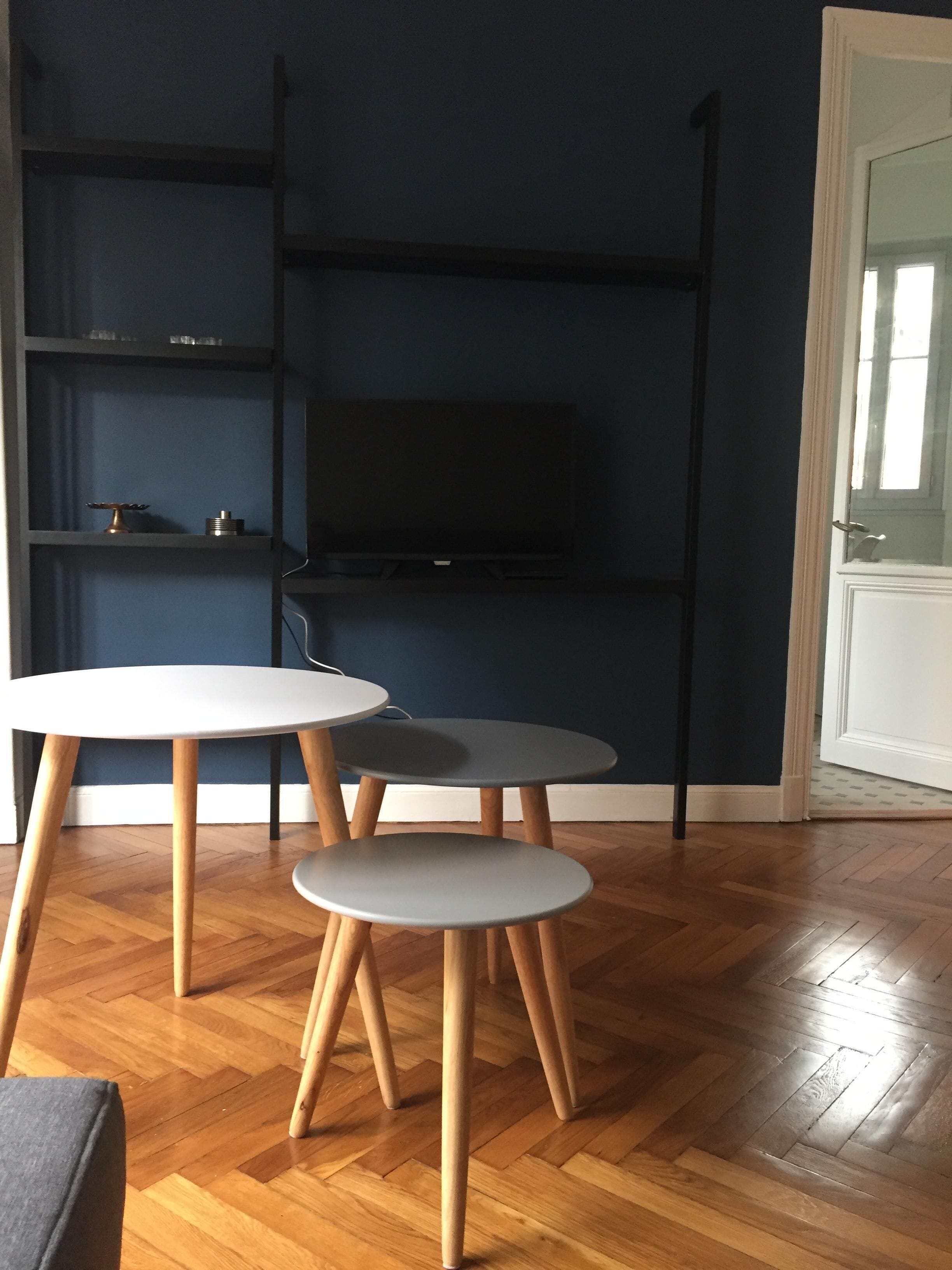 Una habitación con piso de madera en serpeto, una puerta blanca y una pared azul oscuro. Hay tres mesas redondas con patas de madera y un televisor de pantalla plana montado en la pared.