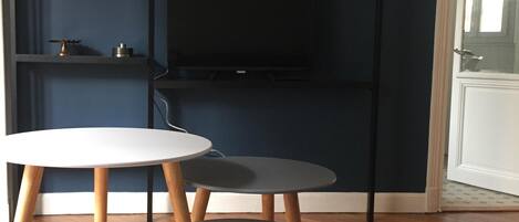 Una habitación con piso de madera en serpeto, una puerta blanca y una pared azul oscuro. Hay tres mesas redondas con patas de madera y un televisor de pantalla plana montado en la pared.