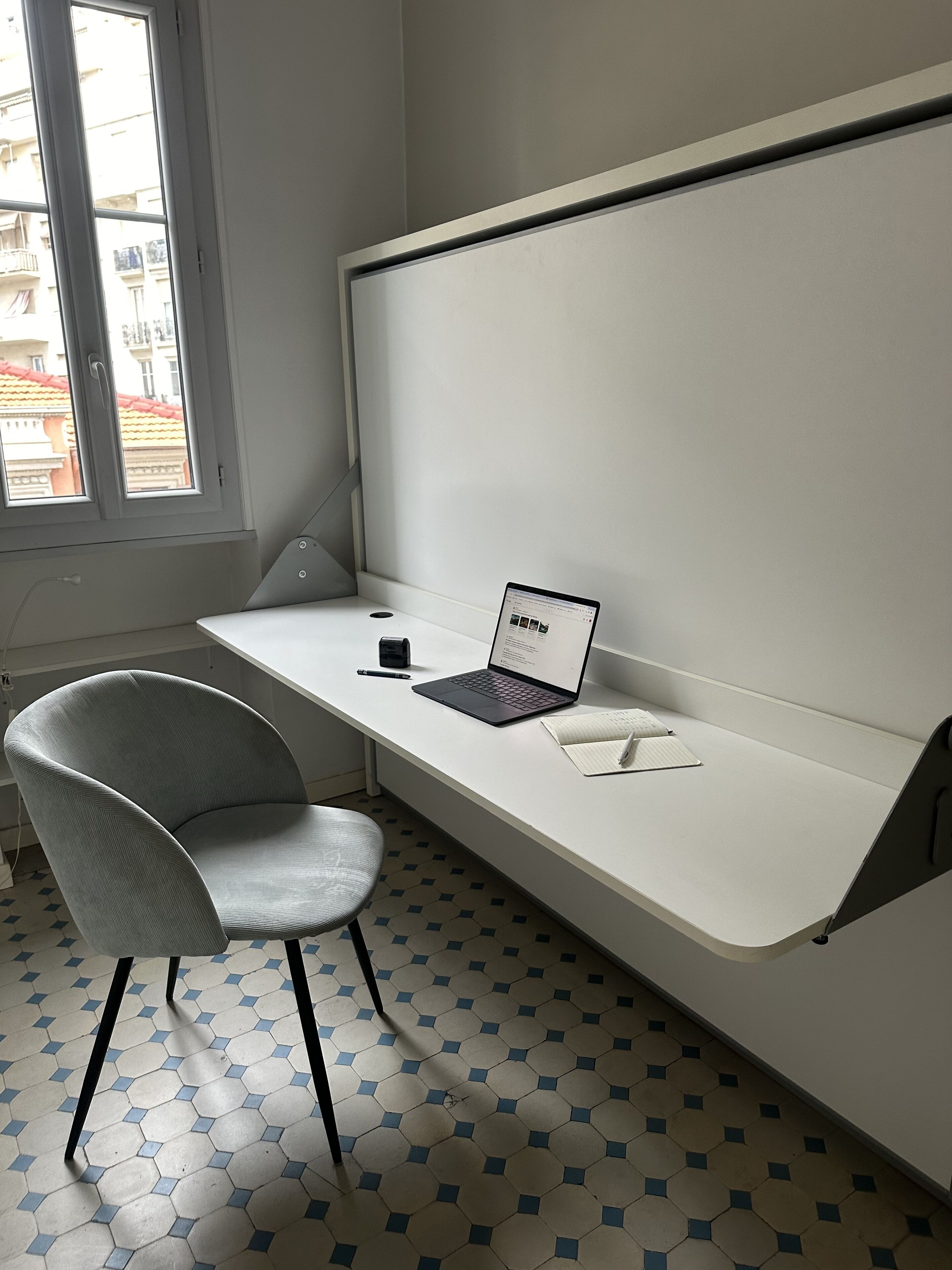 A modern workspace with a white desk, a laptop, a chair, and a window with a view of buildings.