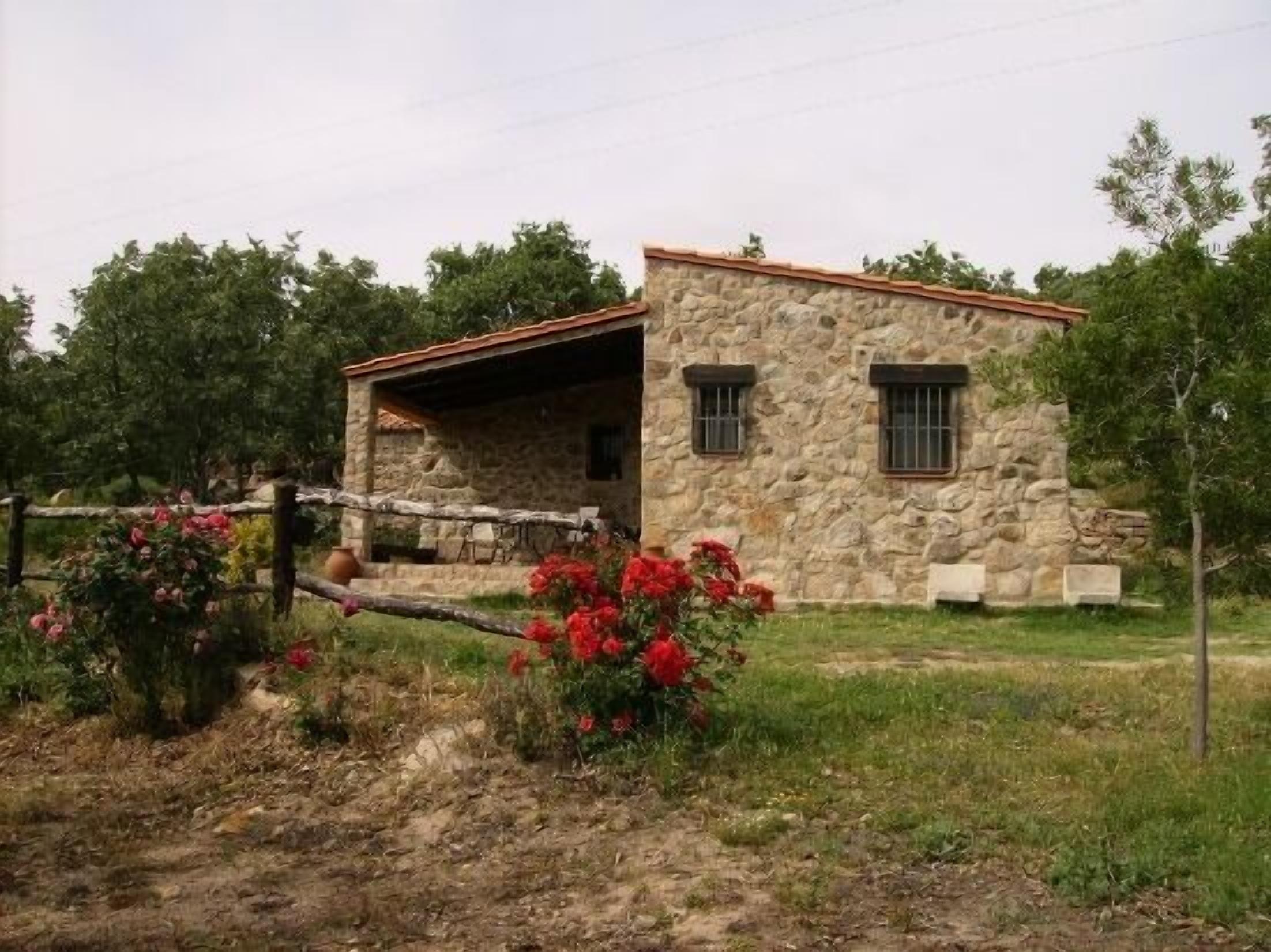 Un bâtiment en pierre avec un toit recouvert de tuiles rouges, un petit porche et une zone délimitée par une clôture, où poussent des fleurs rouges.