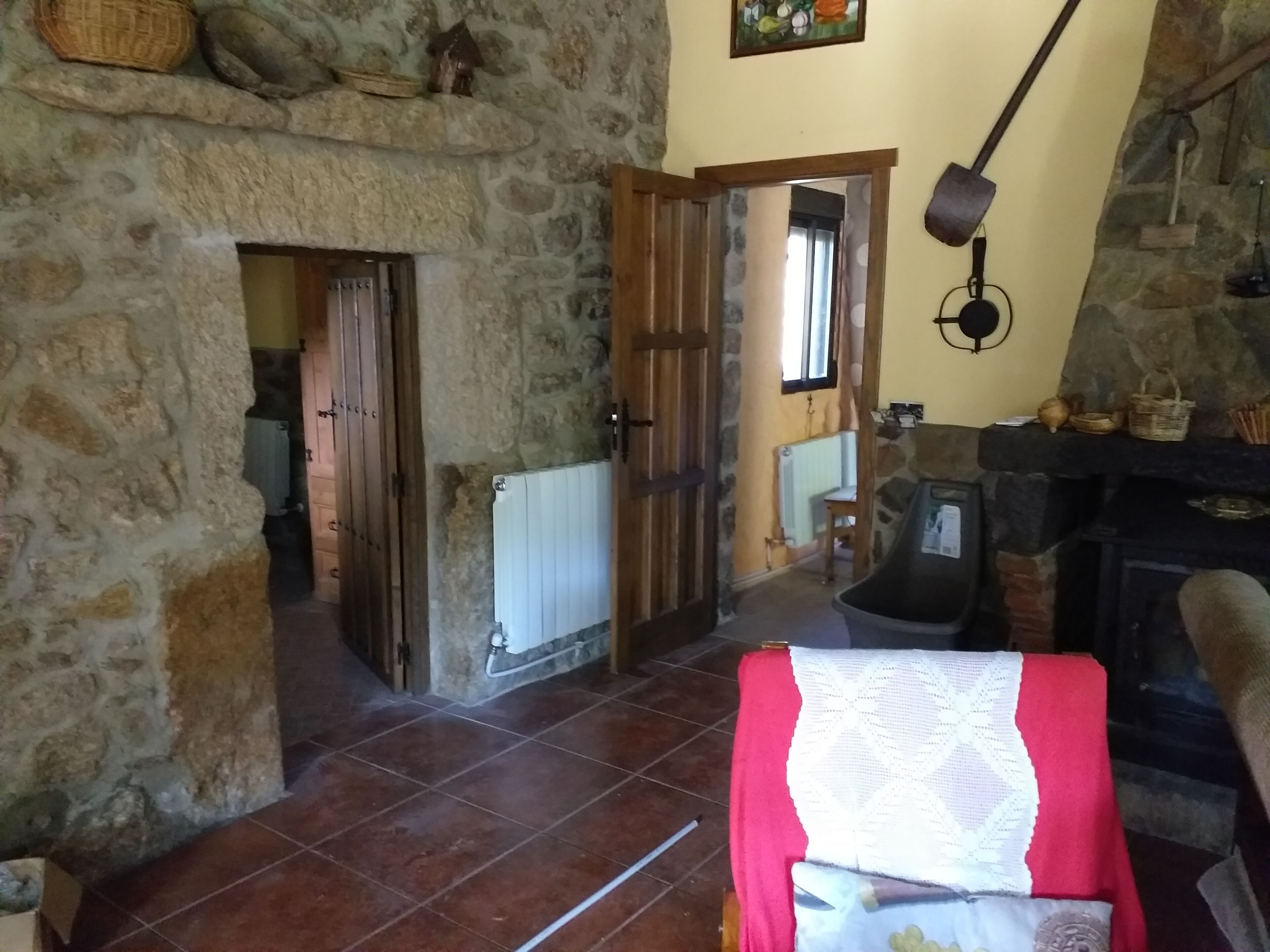 Un intérieur rustique en pierre, avec une porte en bois donnant sur une autre pièce, un sol carrelé et une cheminée sur laquelle est suspendue une marmite.