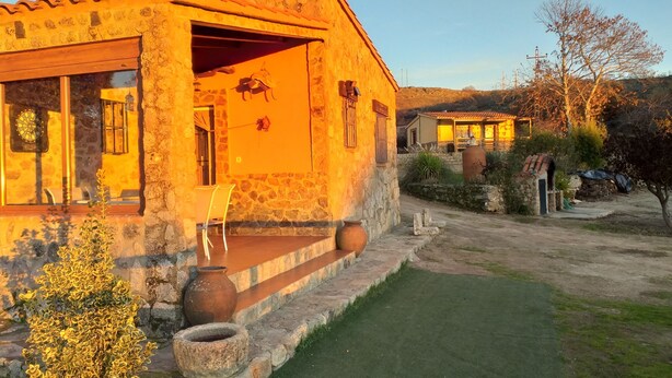 A stone building with a tiled roof, a porch with chairs, and a small garden area.