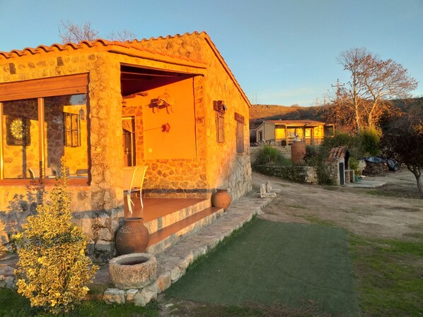 Un edifici de pedra amb teulada de rajola, un porxo amb cadires i una petita zona de jardí.