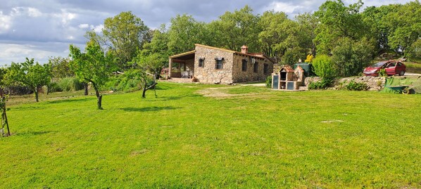 Una casa de piedra con un porche cubierto, un pequeño jardín y un coche rojo aparcado cerca.