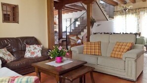 Ein gemütliches Wohnzimmer mit einem braunen Ledersofa, einem beigefarbenen Sofa mit Karokissen und einem Holztisch. Im Hintergrund ist eine Treppe zu sehen.