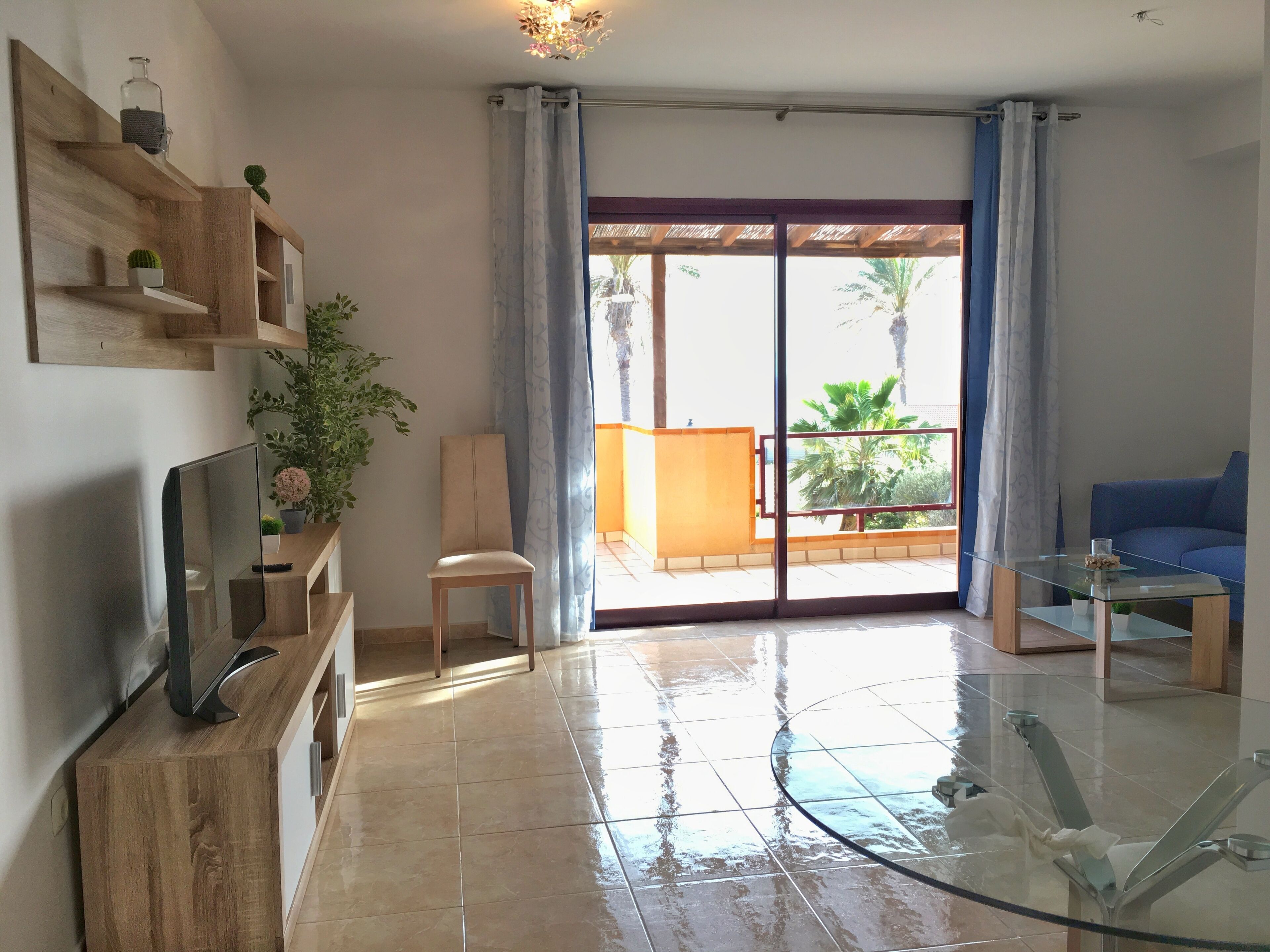 A living room with a TV, glass coffee table, blue sofa, and a sliding glass door leading to a balcony with plants.