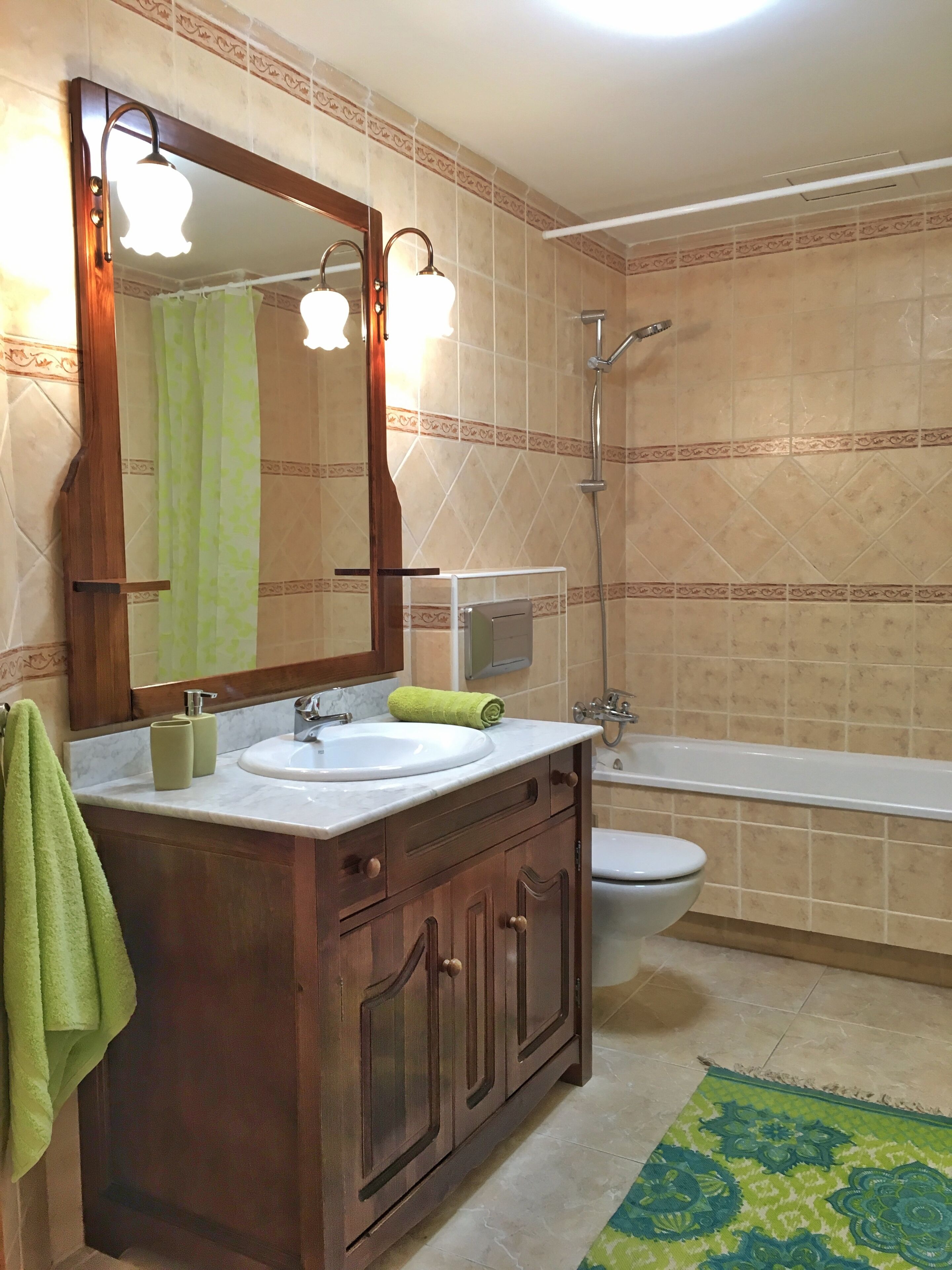 Een badkamer met een houten wastafel, een witte wasbak, een spiegel, een groene handdoek, een douche en een toilet.