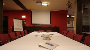 Uma sala de conferências com uma mesa comprida, cadeiras vermelhas, um quadro branco e uma tela de projeção.