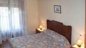 Une chambre à coucher avec un lit, deux tables de chevet avec des lampes, une fenêtre avec des rideaux légers et un tableau encadré au mur.