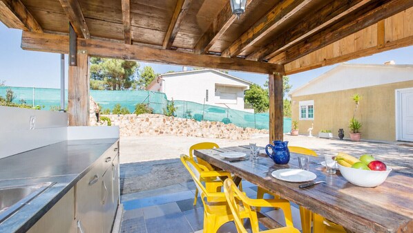 An outdoor patio with a wooden table and yellow chairs, a blue pitcher, and a bowl of fruit.
