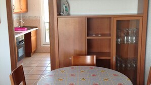 A dining area with a round table covered by a floral tablecloth, surrounded by wooden chairs with blue cushions. A wooden cabinet with glass doors is visible in the background, containing glassware. - Panoramic apartment in Sports & Golf Resort (Sant Jordi)