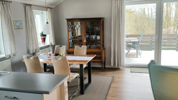 Eine moderne Küche mit einer zentralen Kochinsel, einem Essbereich mit Holztisch und -stühlen, einem Schrank mit Glastüren und Blick durch große Fenster.