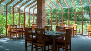 A conservatory with a brick column, wooden tables, and chairs, surrounded by glass walls offering a view of a lush garden. - Black Spur Inn (Narbethong)