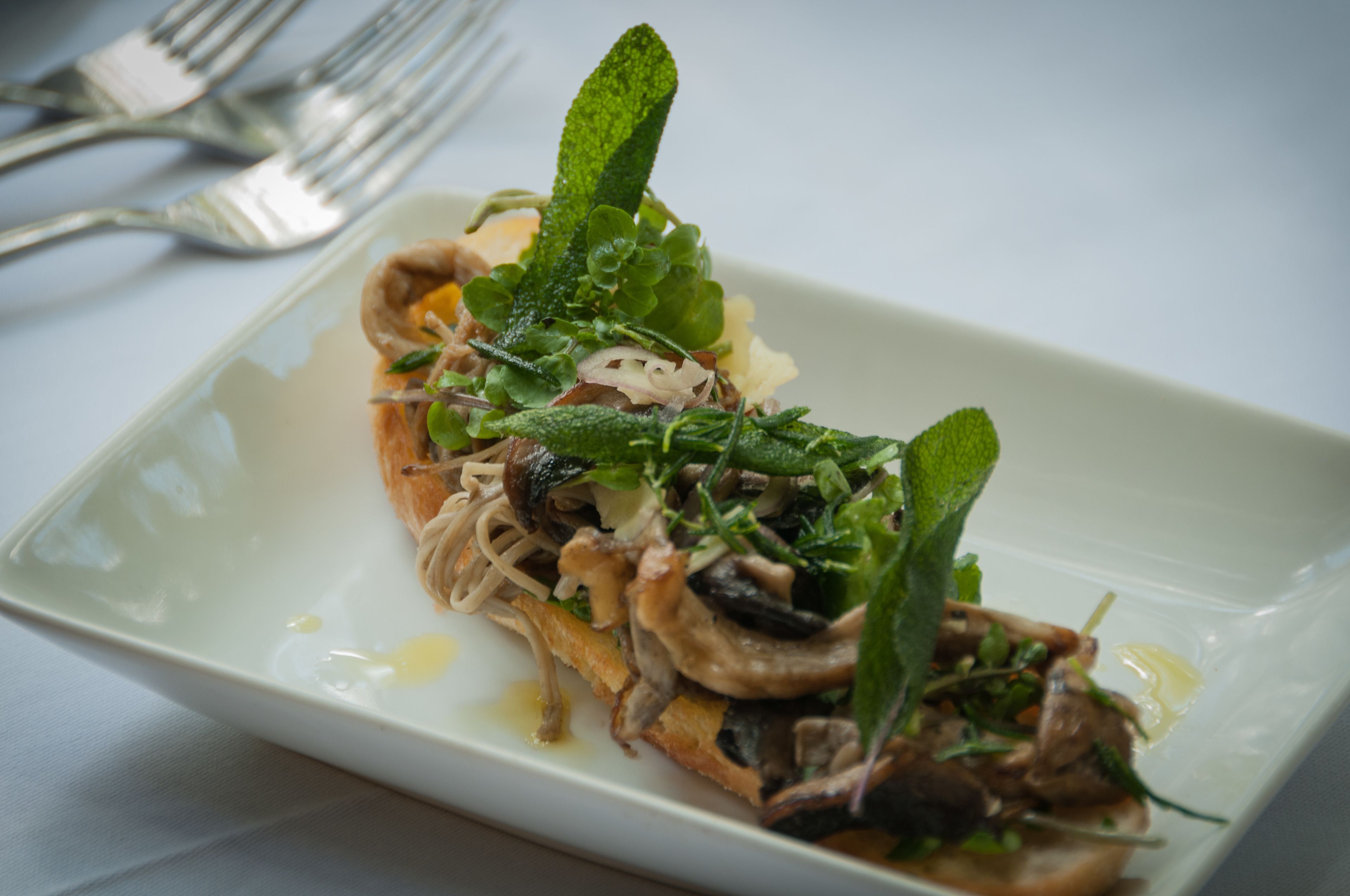 A rectangular plate with a dish of noodles, mushrooms, and greens, accompanied by a fork and knife on a white tablecloth.