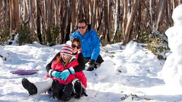 A snowy outdoor scene with a person in a blue jacket and another in a red jacket sitting on the snow.