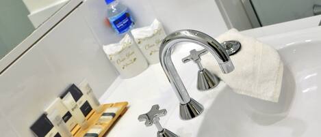 Un baño con un lavamanos blanco, grifo cromado y una botella de agua. También hay dos botellas pequeñas y una bandeja de madera con jabones.