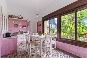 Une cuisine avec un coin repas, des murs roses, un sol carrelé et de grandes fenêtres donnant sur un paysage verdoyant.
