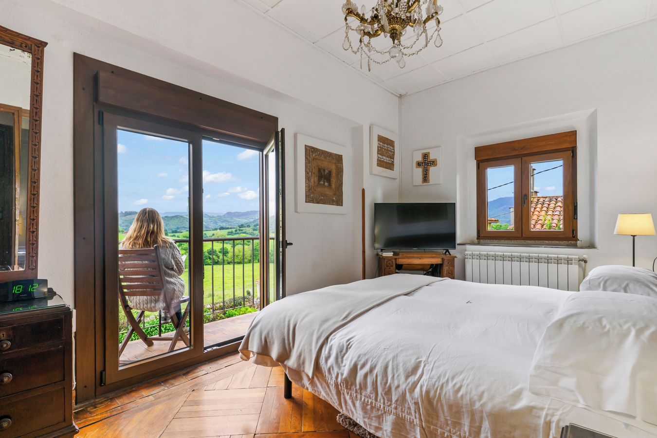A bedroom with a large bed, a television, a window with a view, and a chair by the sliding glass door.