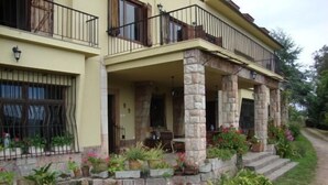 Una casa de dos pisos con balcón, columnas de piedra y un jardín con plantas en macetas.