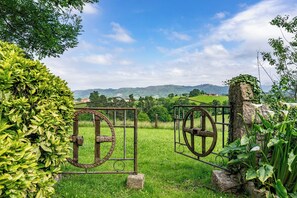Une porte en métal rustique, dotée d’un mécanisme à roue, entourée d’une végétation luxuriante et offrant, en arrière-plan, un paysage pittoresque.