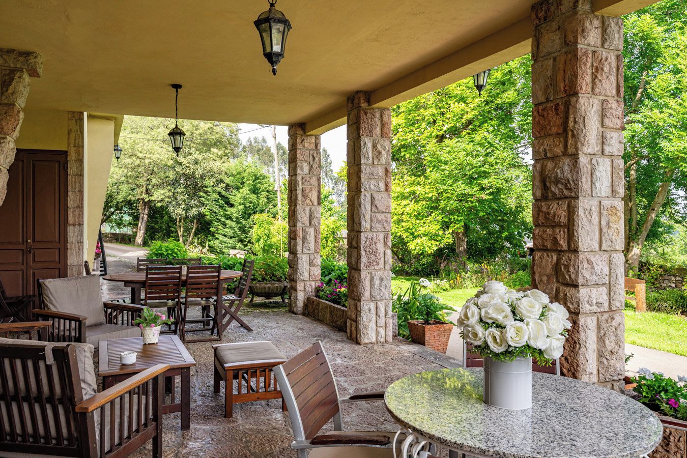 Un espace extérieur aménagé avec des tables et des chaises, protégé par un toit et entouré de verdure, avec des piliers en pierre.