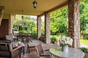 Un espacio de estar al aire libre cubierto, con pilares de piedra, mesas y sillas, rodeado de vegetación.