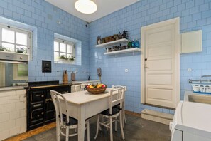 Une cuisine avec des carreaux bleus, une table blanche avec des chaises, un fourneau et une fenêtre ornée de plantes.