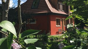Een rode woning met een schuin dak, een veranda en een tuin vol planten en bloemen.