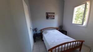 Ein Schlafzimmer mit einem hölzernen Bett, einem Nachttisch, einer Lampe, einem Fenster mit Blick auf Bäume und einem Wandbild.