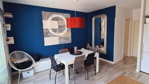 Ein modernes Wohnzimmer mit blauer Akzentwand, einem Essbereich mit weißem Tisch und schwarzen Stühlen, einer roten Pendelleuchte und einem großen Spiegel.