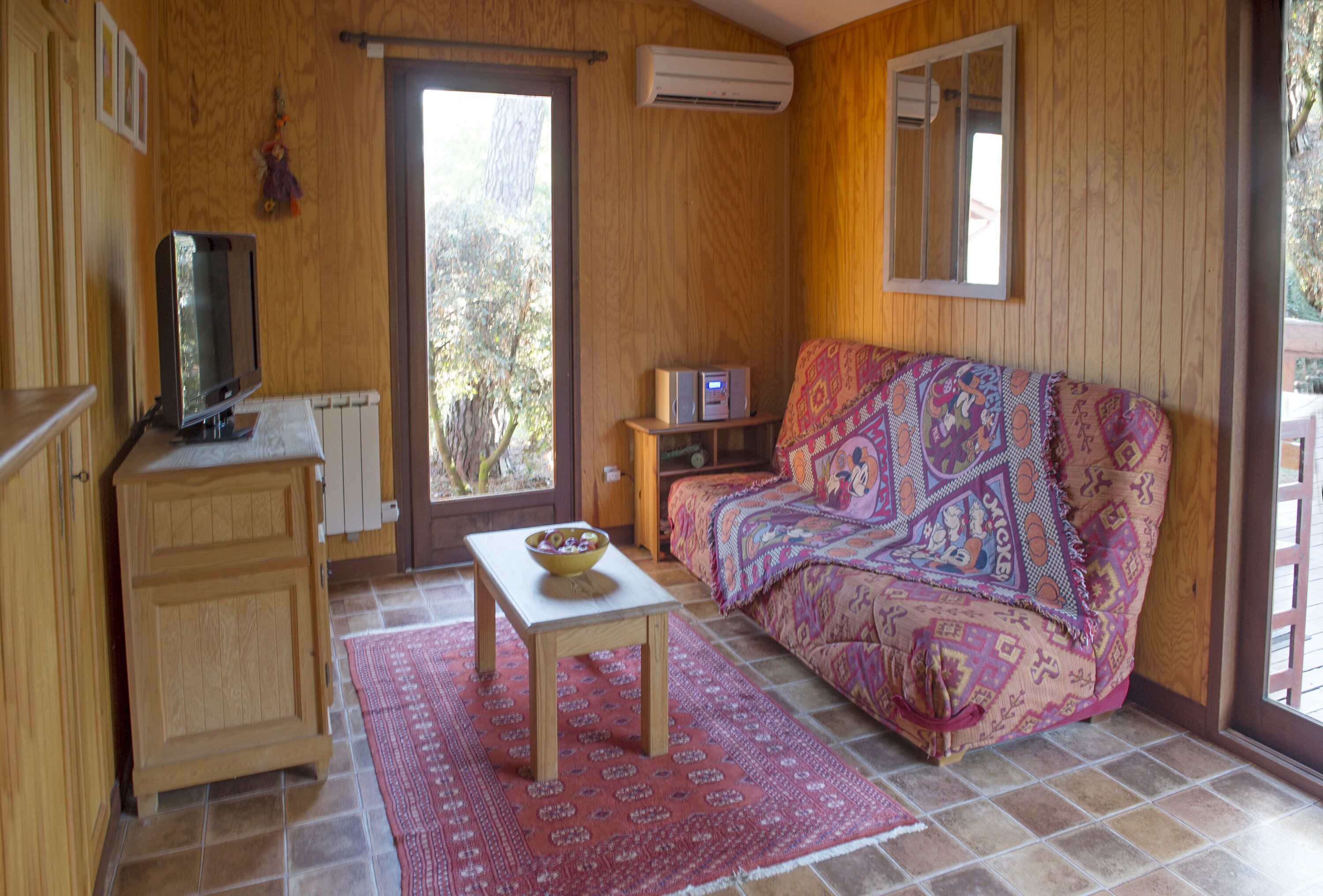 A cozy room with a wooden cabinet, a television, a small table with a bowl, a colorful sofa, and a window with a view of trees.
