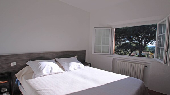 Une chambre à coucher avec un lit, deux oreillers, une table de chevet, une fenêtre donnant sur des arbres et un radiateur.