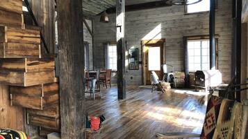 A rustic cabin interior with a wooden staircase, a dining area, and a large window allowing natural light.