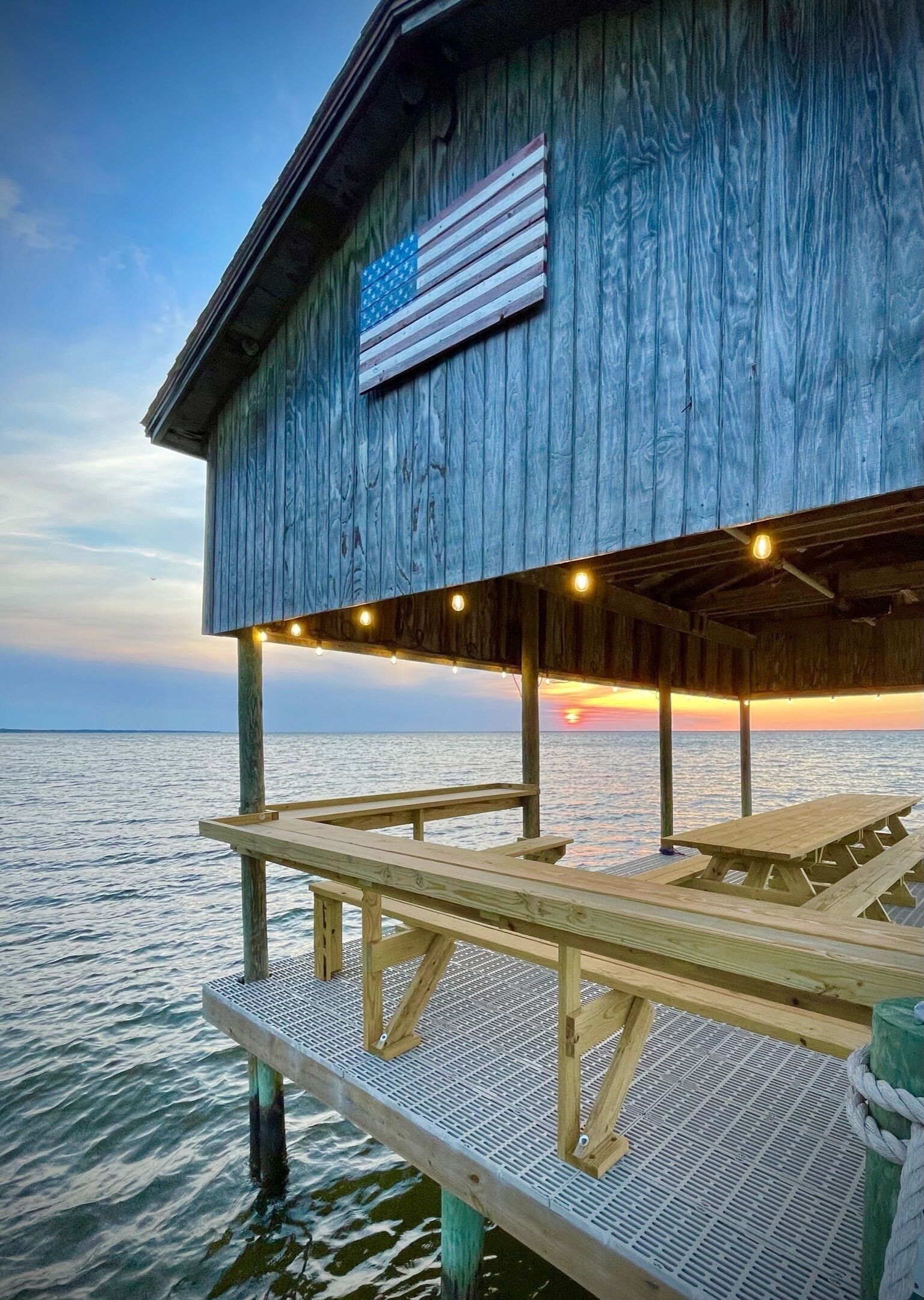 A wooden pier extending over calm water, with a covered seating area and a flag on the building.